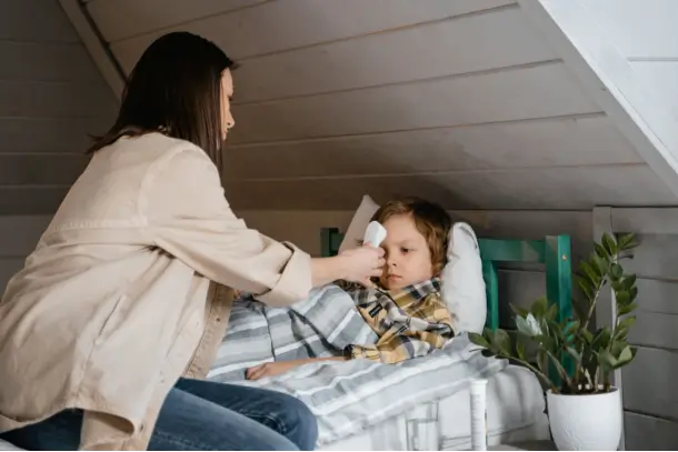 A mother uses a forehead thermometer to check her child’s temperature while the child rests in bed.