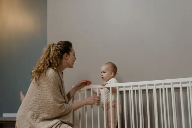 Mother talking to and comforting her baby while the child stands inside a crib