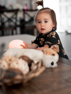 Toddler girl standing against wood table holding an orange VAVA baby night light next to a stuffed toy