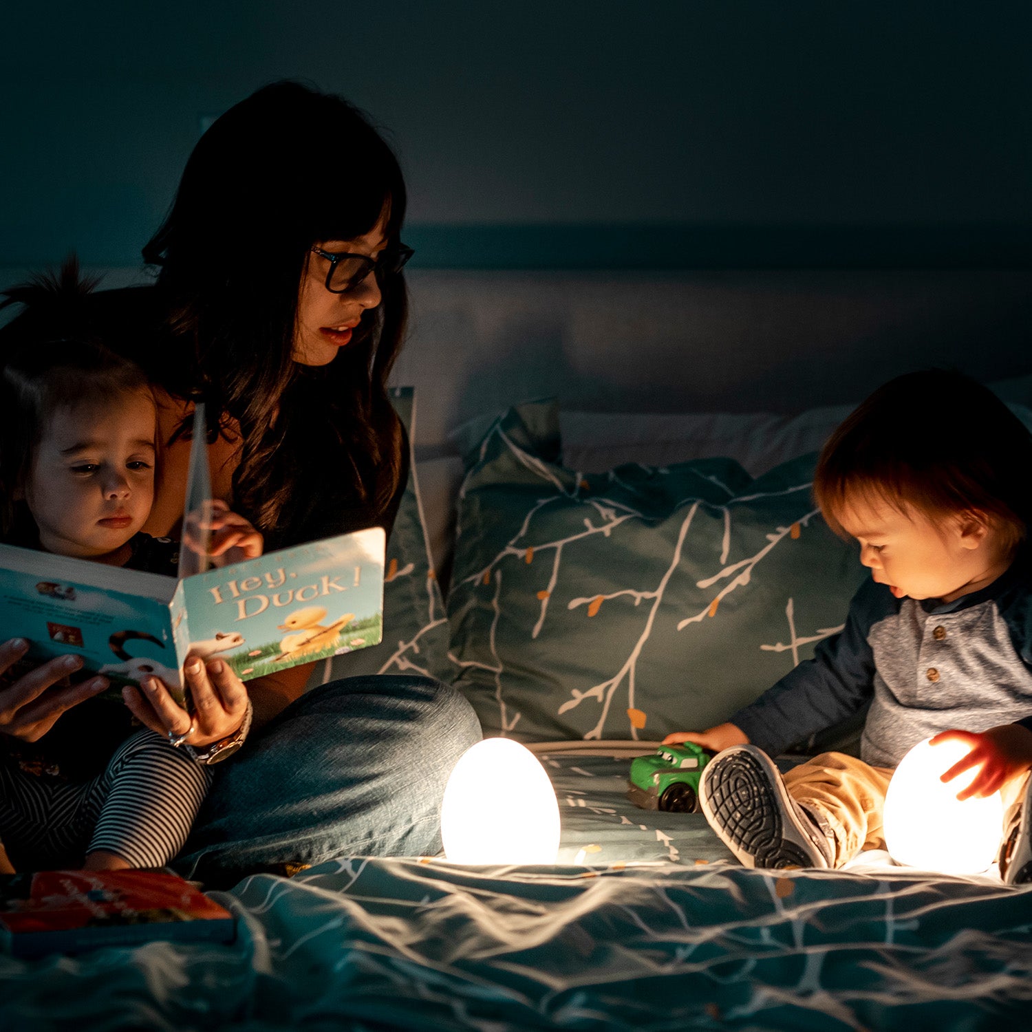 Toddler girl reading a book on a caregiver’s lap next to a VAVA baby night light, toddler boy playing with a green toy car while holding a VAVA baby night light