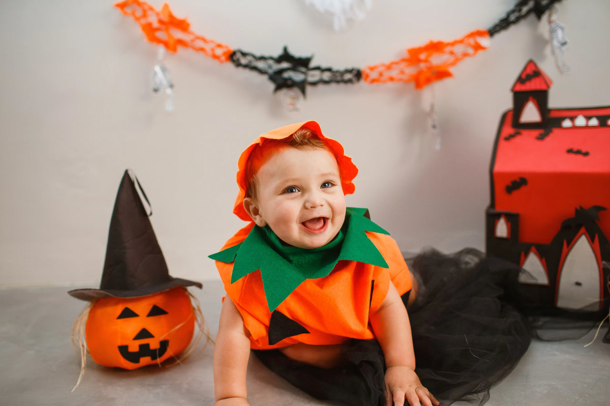 A smiling baby dressed in an orange pumpkin Halloween costume surrounded by festive decorations.