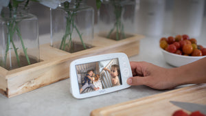 Caregiver holding a VAVA split screen baby monitor on a kitchen counter with bowl of tomatoes in background