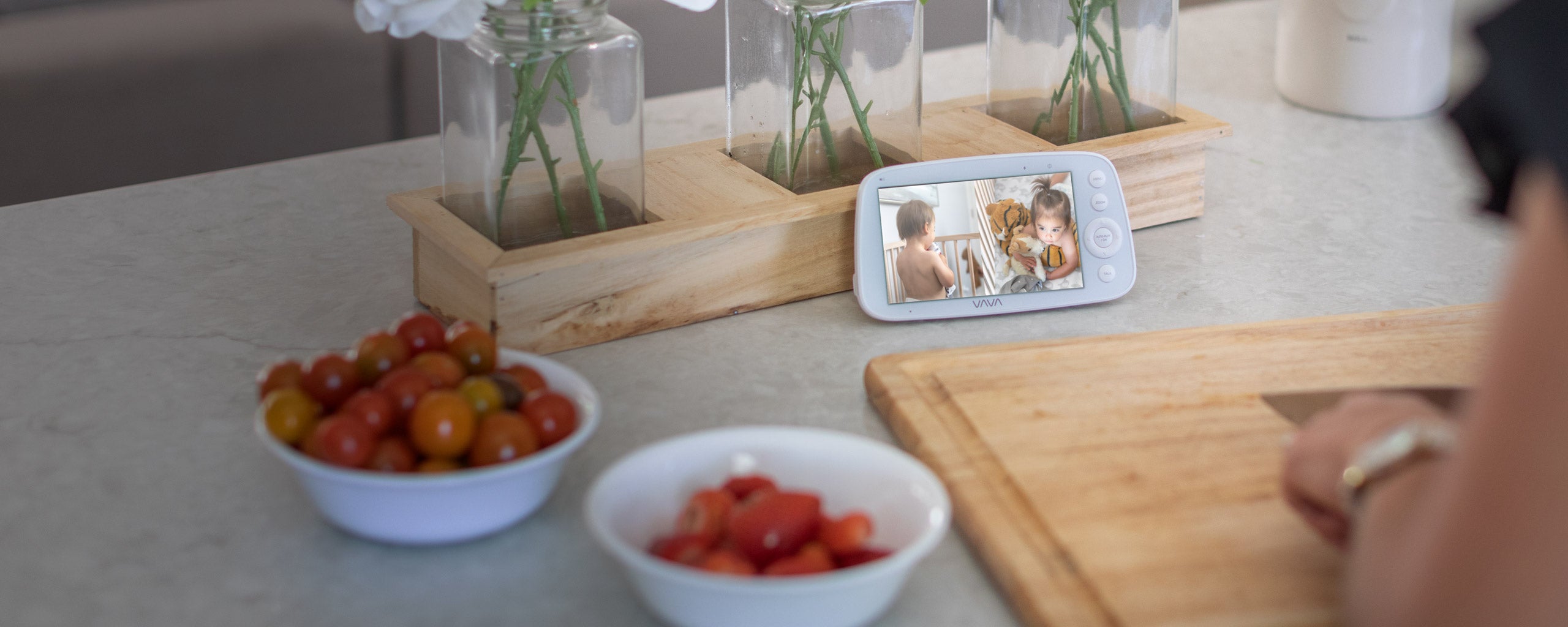 VAVA split screen baby monitor resting on a counter with bowl of tomatoes and brown cutting board in foreground