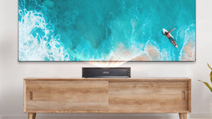 VAVA Chroma Triple Laser 4k Projector on a wood TV Stand with a blue beach on the large screen