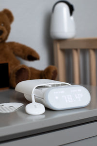 VAVA smart baby thermometer, silicone wand, two adhesive patches on a gray table with a white vase, clock, brown teddy bear, and wood crib railing in the background