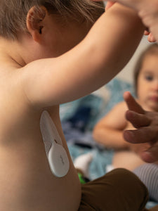 VAVA smart baby thermometer silicone wand and adhesive patch on a child next to a caregivers hand, another child in the background