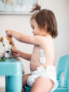 Toddler girl sitting on a blue chair in a diaper wearing a VAVA smart baby thermometer silicone wand and adhesive patch while playing with a stuffed toy dog on a blue table