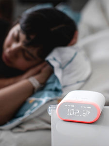 Caregiver sleeping in the background, VAVA smart baby thermometer on a white table displaying 102.3F temperature