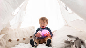 Toddler boy sitting amid white pillows while holding two pink VAVA baby night lights