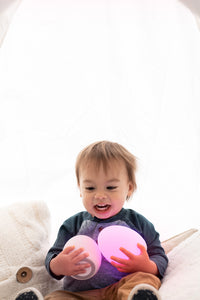 Toddler boy sitting amid white pillows while holding two pink VAVA baby night lights