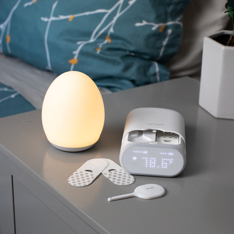 VAVA smart baby thermometer, silicone wand, two adhesive patches and a VAVA night light on a gray table with a turquoise pillow in the background