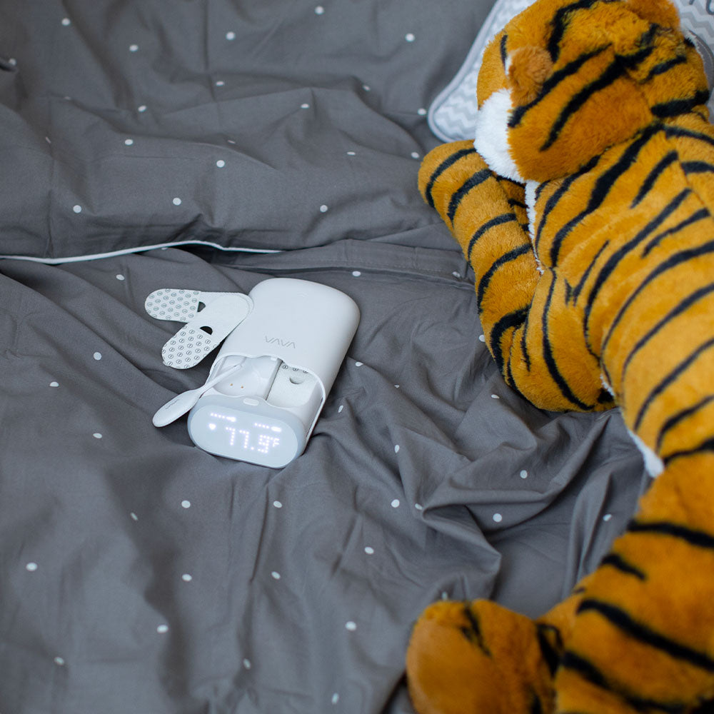 VAVA smart baby thermometer, silicone wand, two adhesive patches on a gray and white polka dot blanket with a stuffed tiger toy
