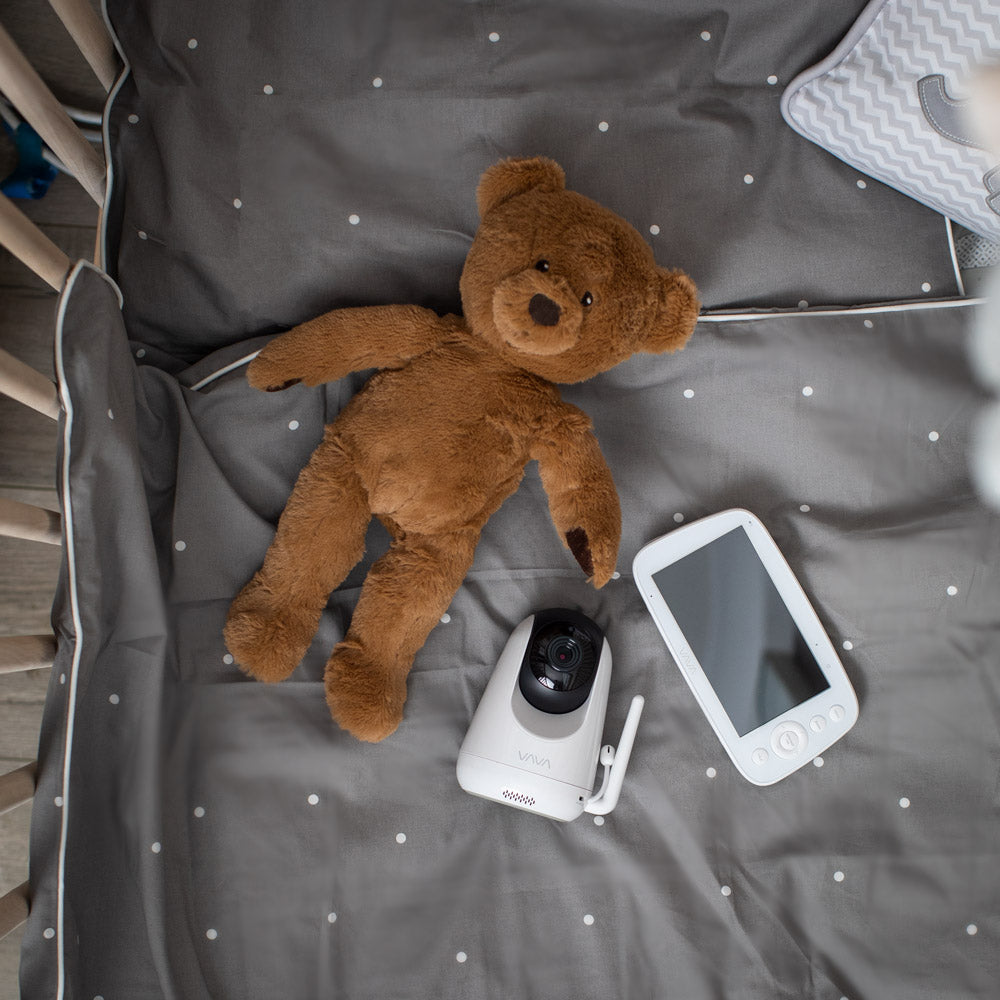 VAVA split screen baby monitor and camera and brown teddy bear in a crib atop a gray blanket with white polka dots
