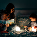 Toddler girl reading a book on a caregiver’s lap next to a VAVA baby night light, toddler boy playing with a green toy car while holding a VAVA baby night light