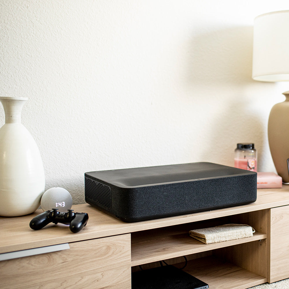 The VAVA 4K Laser TV on a wooden TV stand next to a lamp, candle, vase, and game controller